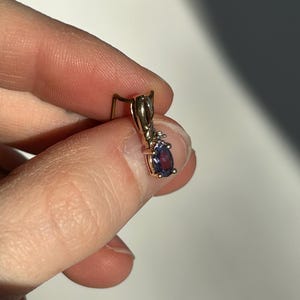 Pode incluir: Um brinco pendente de ouro com uma pedra preciosa roxa escura em forma de gota. O brinco tem uma pequena pedra transparente acima da pedra roxa maior. O brinco &eacute; preso por um gancho de ouro.