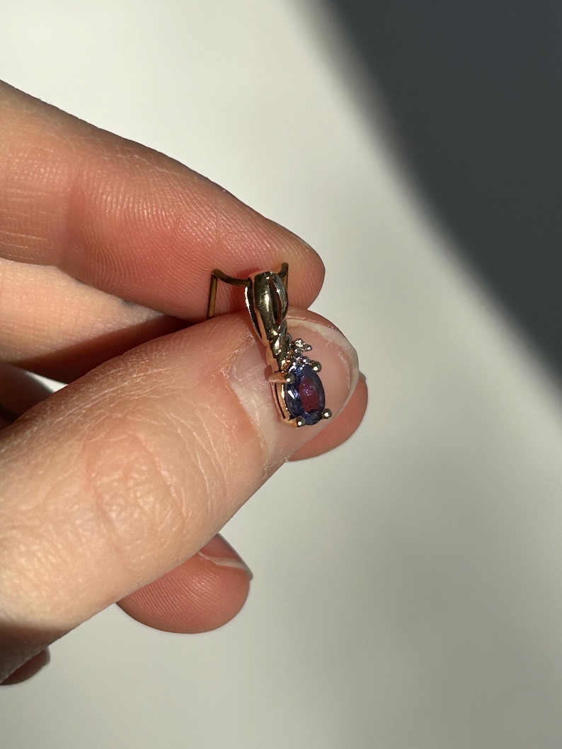 Pode incluir: Um brinco pendente de ouro com uma pedra preciosa roxa escura em forma de gota. O brinco tem uma pequena pedra transparente acima da pedra roxa maior. O brinco &eacute; preso por um gancho de ouro.