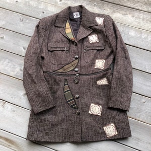 Op de afbeelding: Een bruine tweed blazer met een uniek ontwerp met meerdere klokvormige patches en een ritszak. De blazer heeft een knoopsluiting en een kraag.