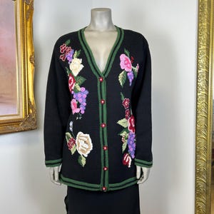 Könnte beinhalten: Schwarze Strickjacke mit floraler Stickerei. Der V-Ausschnitt-Pullover hat grüne Verzierungen und rote Knöpfe. Das florale Design umfasst rosa, lila und weiße Blumen mit grünen Blättern. Das Kleidungsstück wird auf einer Schaufensterpuppe präsentiert.