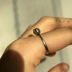 K&ouml;nnte beinhalten: Ein silberner Ring mit einem einzigen runden Edelstein, der in einer Krappenfassung sitzt.