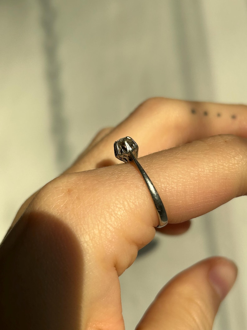 K&ouml;nnte beinhalten: Ein silberner Ring mit einem einzigen runden Edelstein, der in einer Krappenfassung sitzt.
