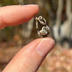 May include: A small, gold-colored pendant with a heart-shaped design. The pendant is held between two fingers against a blurred background of trees. The heart is centered within a teardrop-shaped frame, and the pendant has a small loop at the top for a chain.