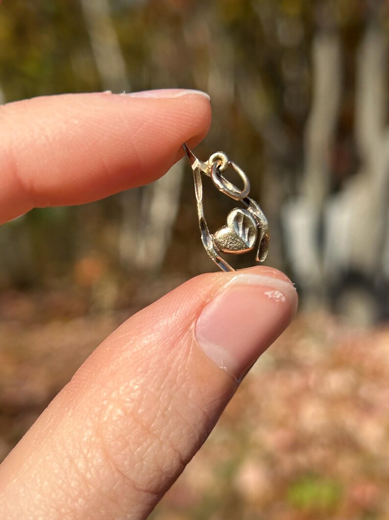 May include: A small, gold-colored pendant with a heart-shaped design. The pendant is held between two fingers against a blurred background of trees. The heart is centered within a teardrop-shaped frame, and the pendant has a small loop at the top for a chain.
