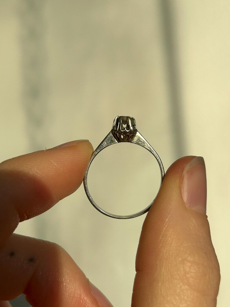 K&ouml;nnte beinhalten: Ein silberner Ring mit einem einzigen runden Edelstein, der in einer Krappenfassung sitzt.
