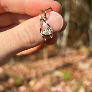 May include: A gold-colored pendant with a heart-shaped charm. The pendant has a delicate, open design with a textured heart at the center. The pendant is held up by a finger.