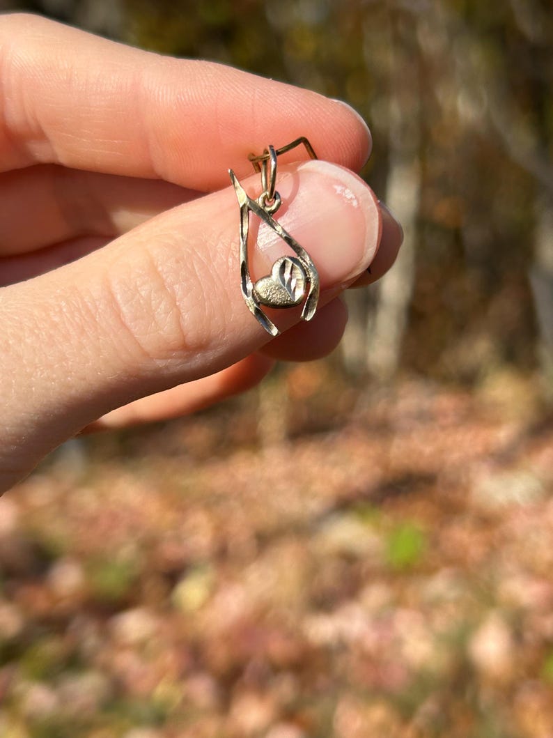 May include: A gold-colored pendant with a heart-shaped charm. The pendant has a delicate, open design with a textured heart at the center. The pendant is held up by a finger.