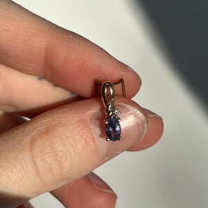 Pode incluir: Um pingente de ouro com uma pequena pedra preciosa oval azul escuro e um pequeno diamante. O pingente &eacute; segurado entre os dedos contra um fundo neutro. &Eacute; uma joia.