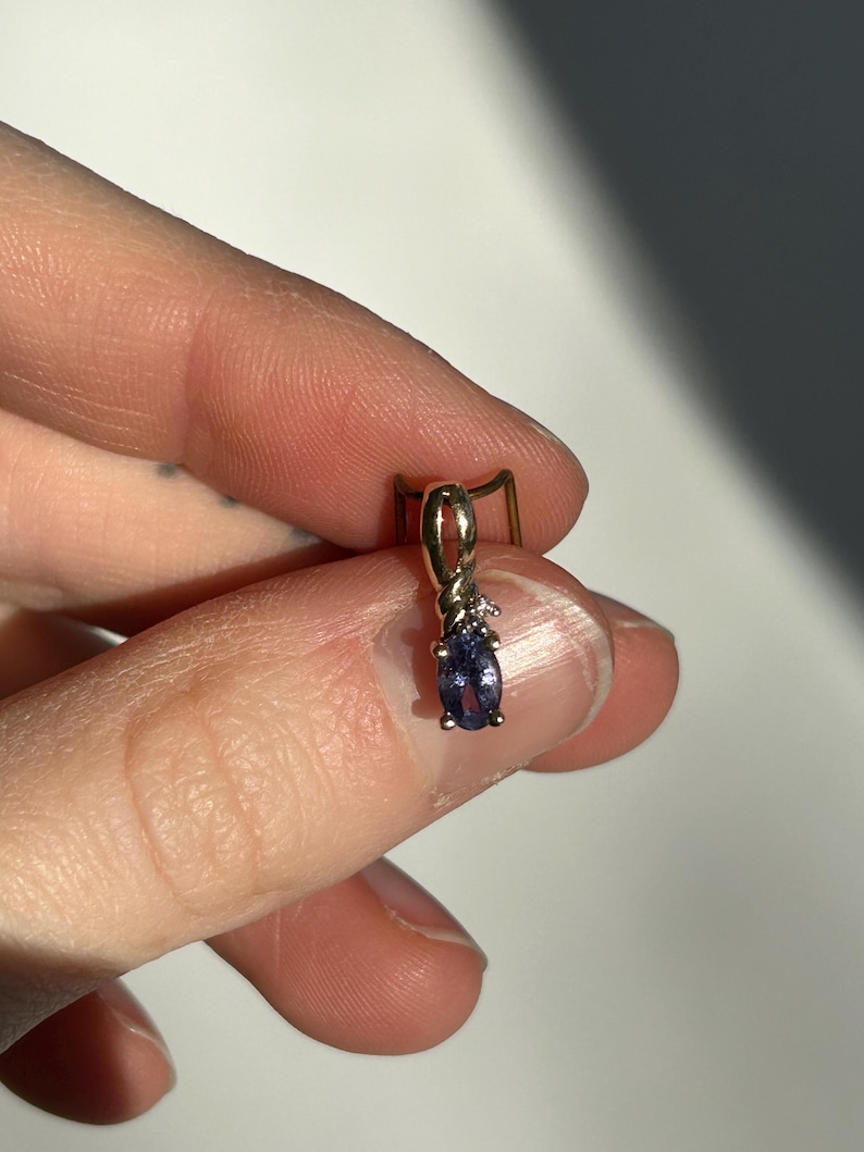 Pode incluir: Um pingente de ouro com uma pequena pedra preciosa oval azul escuro e um pequeno diamante. O pingente &eacute; segurado entre os dedos contra um fundo neutro. &Eacute; uma joia.