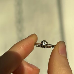 K&ouml;nnte beinhalten: Ein silberner Ring mit einem einzigen, runden, klaren Edelstein, der in einer Krappenfassung sitzt.