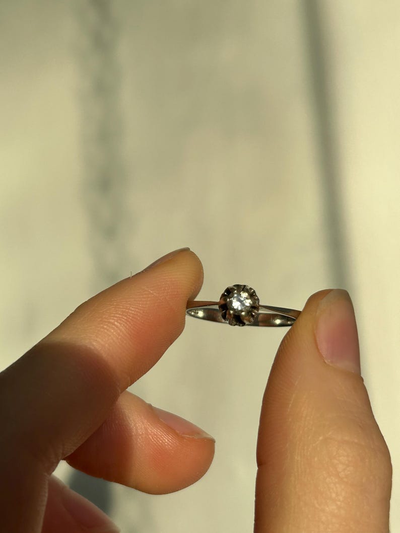 K&ouml;nnte beinhalten: Ein silberner Ring mit einem einzigen, runden, klaren Edelstein, der in einer Krappenfassung sitzt.