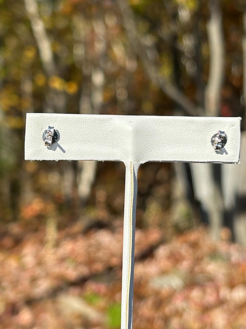 K&ouml;nnte beinhalten: Ein Paar silberne Ohrstecker mit einem Cluster aus kleinen, klaren Steinen. Die Ohrringe werden auf einer wei&szlig;en Ohrringkarte vor einem verschwommenen Hintergrund aus Herbstlaub pr&auml;sentiert.