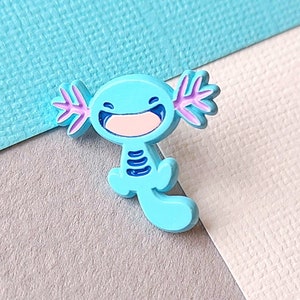 May include: A blue axolotl enamel pin with purple gills and a pink smile. The pin is on a light blue and grey background.