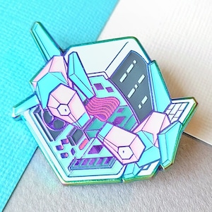 May include: A teal, pink and purple enamel pin of a robot with a keyboard and a screen. The robot is in a sitting position with its legs crossed.