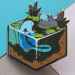 Puede incluir: Un ajolote azul y morado con un ajolote más pequeño en una roca en un cubo transparente con nenúfares verdes y tierra marrón. El cubo está rodeado por un marco de metal plateado.