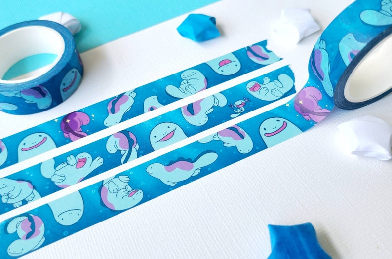 May include: Blue washi tape with a repeating pattern of light blue and purple sea creatures on a dark blue background. The tape is rolled up on a white surface.