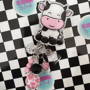 Puede incluir: Un portatarjetas con temática de vaca con un diseño de vaca de dibujos animados. El portatarjetas presenta una vaca de plástico transparente con manchas blancas y negras, detalles en rosa y purpurina. El dije incluye cuentas rosas y negras, y la palabra "MOO". El fondo es un patrón de cuadros blancos y negros.