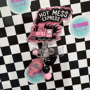 以下が含まれることがあります： 「HOT MESS EXPRESS」の文字と列車のデザインが施されたピンクと黒のバッジリール。 「ALL ABOARD！」の文字とその他の装飾要素が付いたチャームが含まれています。 背景は白と黒のチェック柄です。