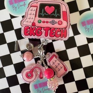 May include: A pink glittery badge reel featuring an EKG monitor graphic with a heart rhythm and the text EKG TECH. Attached are charms including a pink stethoscope, a heart rhythm strip, and two pink beads on a silver metal clip.