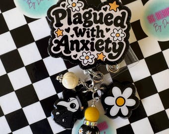 Puede incluir: Un portatarjetas negro con el texto blanco Plagued With Anxiety, decorado con estrellas amarillas y flores blancas. Incluye dijes colgantes: un médico de la peste, una flor blanca, una cuenta amarilla y una cuenta negra con margaritas.