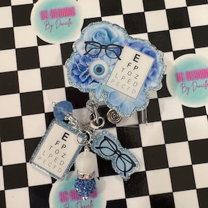 May include: A blue and silver badge reel with an eye chart, eyeglasses, and a blue flower design. The reel includes a charm with a blue crystal design and the text "SG Designs By Danielle".