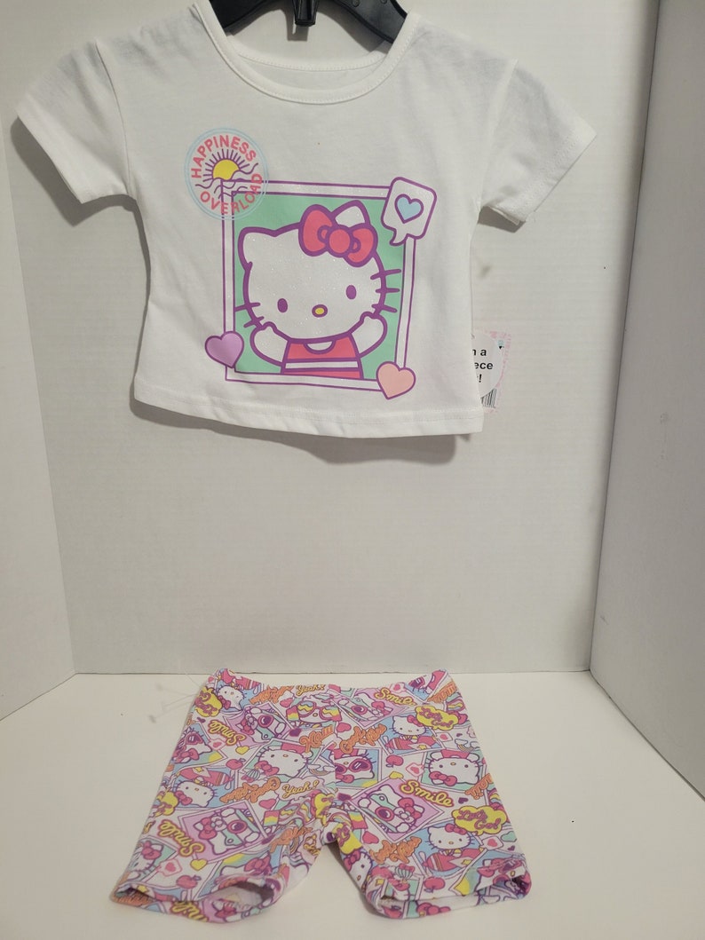May include: A white t-shirt with a Hello Kitty graphic and the text "Happiness Overload". The shirt is paired with pink and white shorts featuring a Hello Kitty print.