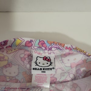 May include: A white Hello Kitty leggings with a pink, blue, and yellow print. The leggings have a tag that says "Hello Kitty" and "18M". The tag also has instructions for care in English, Spanish, and French.