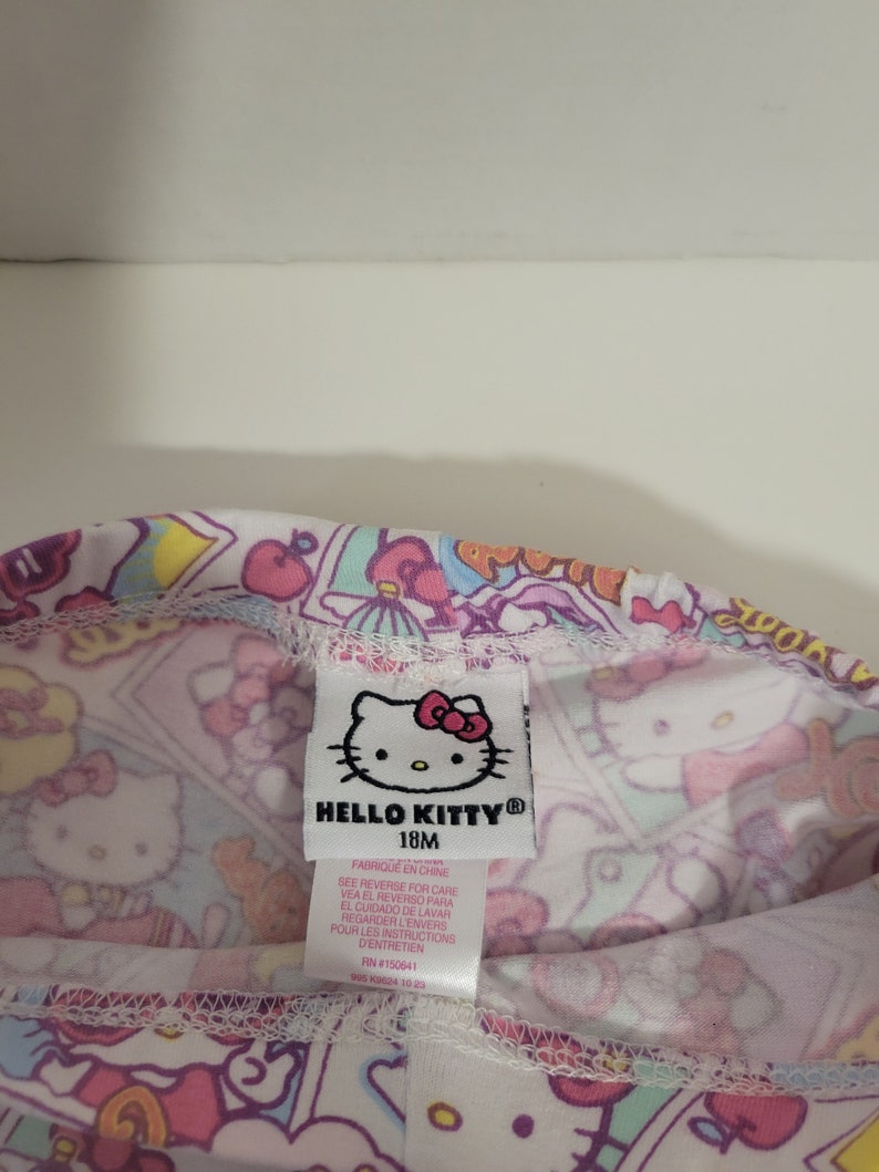 May include: A white Hello Kitty leggings with a pink, blue, and yellow print. The leggings have a tag that says "Hello Kitty" and "18M". The tag also has instructions for care in English, Spanish, and French.