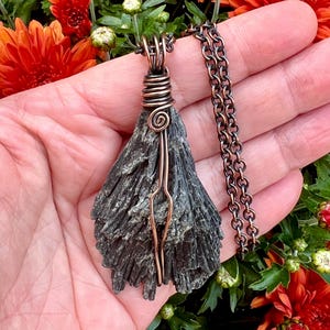May include: A handcrafted necklace featuring a dark gray, textured stone pendant. The pendant is wrapped with copper wire and suspended from a copper-colored chain. The necklace is displayed against a backdrop of orange flowers and green foliage.
