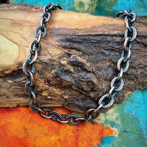 May include: A heavy metal chain necklace featuring oval-shaped links with a dark, oxidized finish and intricate, etched floral patterns on the surface of each individual link, creating a textured and weathered appearance throughout the piece.