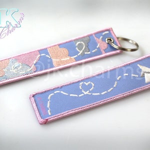 May include: Two pink and blue fabric keychains with embroidered designs. One keychain features a white paper aeroplane with a heart and the other features a series of hearts.