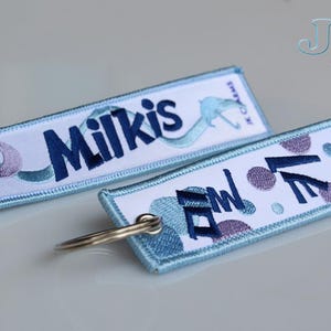 Puede incluir: Dos llaveros rectangulares con texto y diseños bordados. Un llavero dice "Milkis" en azul oscuro, con un diseño azul claro. El otro llavero tiene las palabras "FAN" y "LOVE" en azul oscuro, con acentos morados y azules.