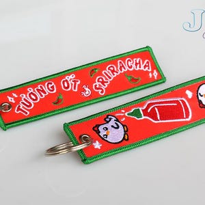 May include: Two red and green keychains with embroidered designs. One keychain features the text "TƯƠNG ỚT & SRIRACHA" and a red chilli pepper. The other keychain features a cartoon chicken and a bottle of red hot sauce.