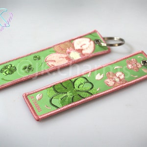 Peut inclure: Deux porte-clés verts et roses avec des trèfles à quatre feuilles et des fleurs brodés. Les porte-clés ont une bordure rose et un anneau de clé argenté.