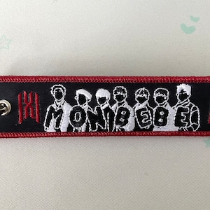 May include: Black and red key chain with a white embroidered image of seven people and the text "MONSTA X" on a black background.