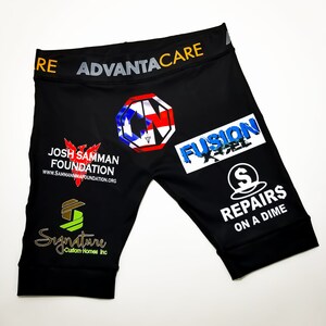 May include: Black athletic shorts with various logos and text printed on them. The logos include "Josh Samman Foundation", "Fusion X-Cel", "Signature Custom Homes Inc", and "Repairs on a Dime". The text "ADVANTACARE" is printed on the waistband.