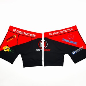 May include: Red and black athletic shorts with the text "ZENKO FIGHTWEAR" and "BIG SIOUX CONSTRUCTION" on the waistband. The shorts also feature the text "NEXT EDGE" and "TEAM HOWE" in white.