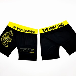 May include: Black athletic shorts with yellow trim and a tiger graphic on the left leg. The shorts have "Zenko Fightwear" and "RMT Fitness" text on the front and "Rad Muay Thai" text on the back.