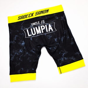 May include: Black athletic shorts with a yellow waistband and trim. The shorts have a white print that says "Uncle J's Lumpia" and "Sadeck Sanon".