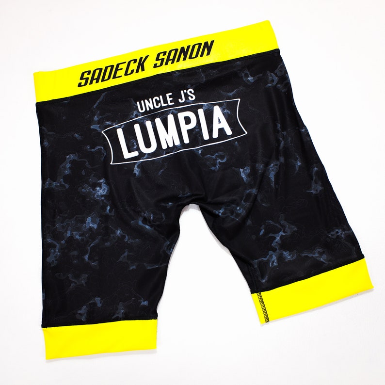 May include: Black athletic shorts with a yellow waistband and trim. The shorts have a white print that says "Uncle J's Lumpia" and "Sadeck Sanon".