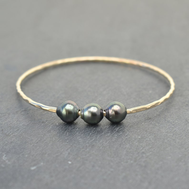 May include: A gold bangle bracelet with three black pearls. The bracelet is hammered and has a simple design.