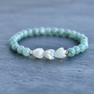 May include: A stretch bracelet featuring round, pale green jade beads and three white, carved mother-of-pearl flower-shaped beads at the center. The floral beads are accented by small, polished gold-tone metal spacers on either side.