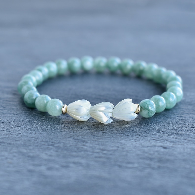 May include: A stretch bracelet featuring round, pale green jade beads and three white, carved mother-of-pearl flower-shaped beads at the center. The floral beads are accented by small, polished gold-tone metal spacers on either side.