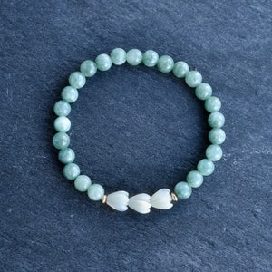 May include: A stretch bracelet composed of smooth, round, light green jade beads. The design features three white, carved flower-shaped beads positioned at the center, accented by small, metallic gold-toned spacer beads on either side of the floral cluster.