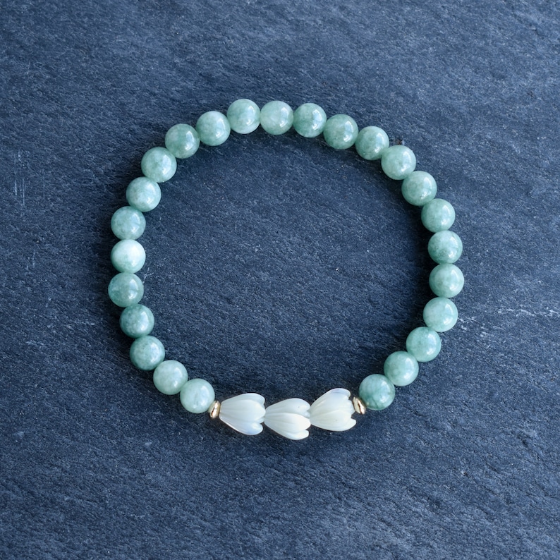May include: A stretch bracelet composed of smooth, round, light green jade beads. The design features three white, carved flower-shaped beads positioned at the center, accented by small, metallic gold-toned spacer beads on either side of the floral cluster.