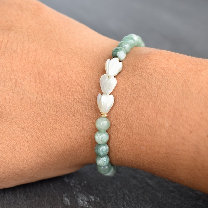May include: A beaded bracelet featuring round, mottled light green stone beads and three white, carved flower-shaped beads arranged in a row. The beads are strung together with a small gold-toned spacer bead separating the stone and flower elements.