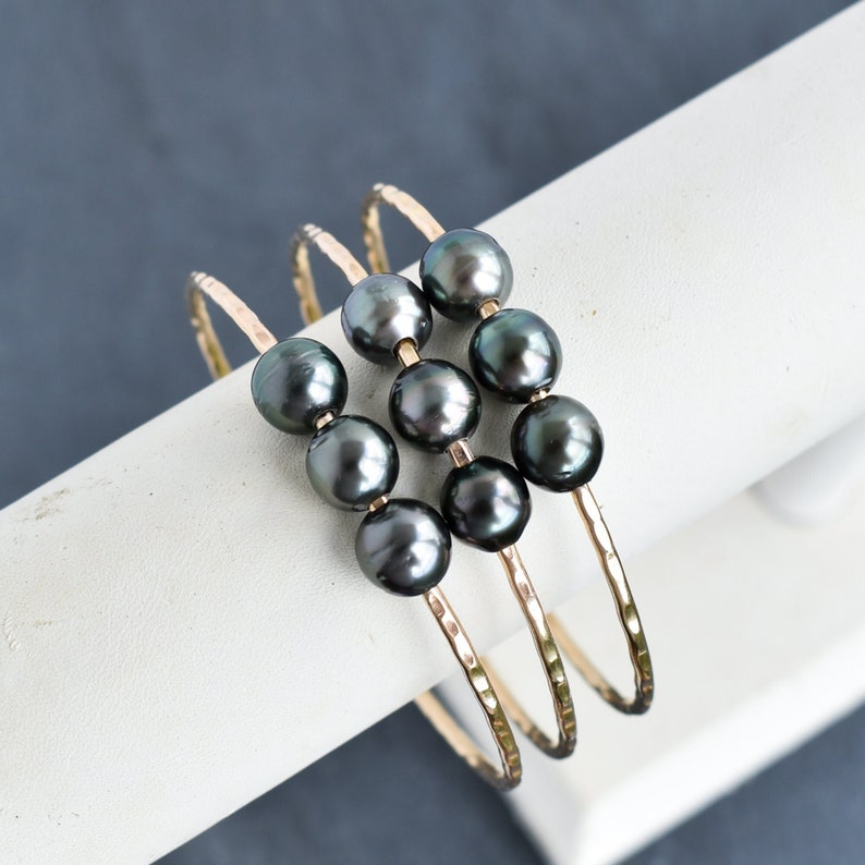 May include: Three gold bangle bracelets with black pearls. The bracelets are stacked on top of each other and have a hammered texture.