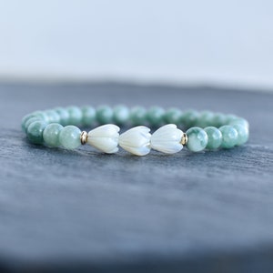 May include: A stretch bracelet featuring smooth, round, light green jade-like beads. The center of the bracelet showcases three white, carved flower-shaped beads made of shell, separated by small, polished gold-tone metal spacer beads.
