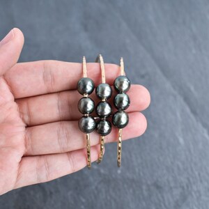 May include: Three gold bangles with black pearls. The bangles are stacked on top of each other and have a hammered texture.