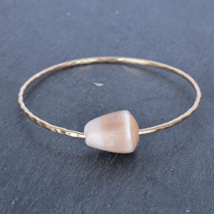 Może przedstawiać: Złota bransoletka typu bangle z młotkowanym wykończeniem i jedną jasnobrązową perłą z muszli.